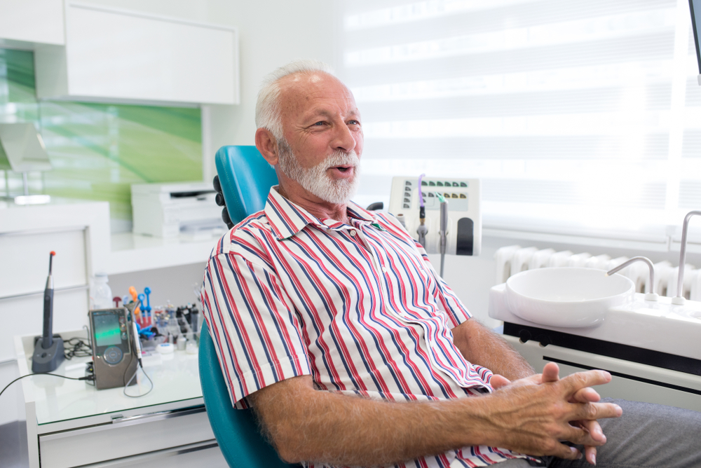 An older man with white hair and a beard sits in a dental chair, wearing a red and white striped shirt. He smiles with hands clasped. The bright clinic room shows dental tools and equipment in the background.