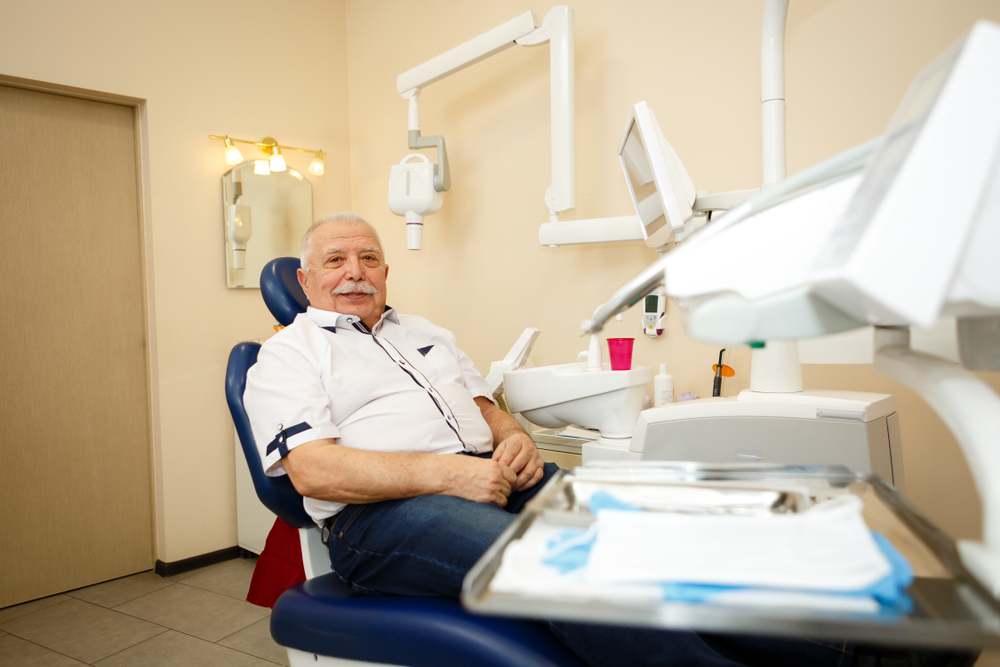 An older man with a mustache sits in a dentist’s chair, wearing a white shirt. Dental equipment and a tray with tools are nearby. The room has beige walls, a mirror, and visible dental machinery.