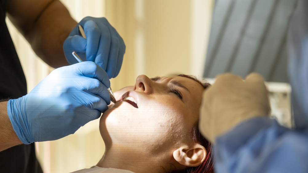 A person reclines in a dental chair as two gloved dentists use instruments to examine their mouth. Sunlight streams in from the left, illuminating the patient’s face. The dentists’ faces are not visible.