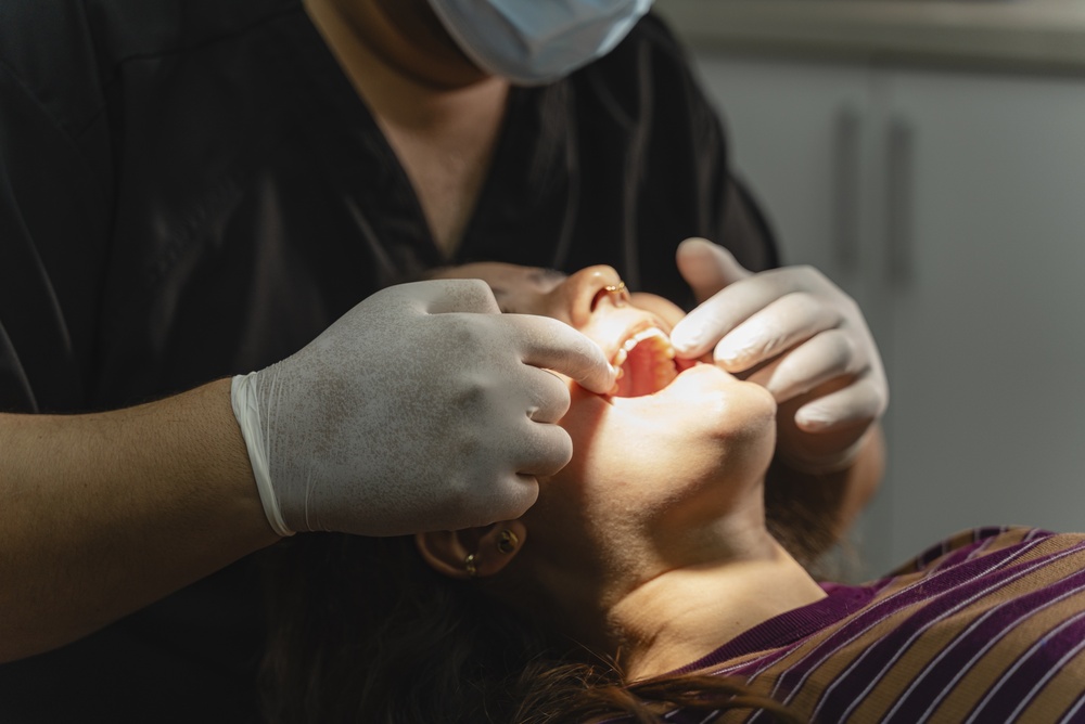 A dentist wearing gloves and a mask examines a patient’s mouth. The patient, in a striped shirt, lies back in the dental chair. The background shows the dental office setting.